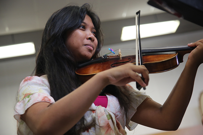A violinist in a white embroidered top playing her instrument with a look of concentration. She has a medium-brown complexion and long dark hair.