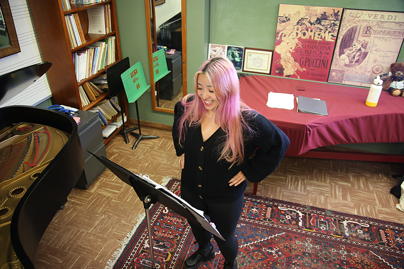 A vocalist standing in front of a poster that reads La Boheme looking at her music stand. She is young and Asian and has long pink hair. She is dressed in a black long-sleeved shirt and black pants.