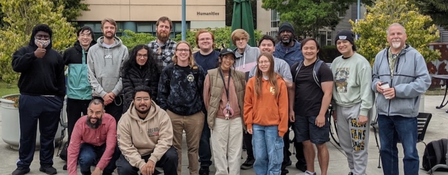 A group of students of diverse ethnicity and gender representation wearing casual clothing stand outside the Creative Arts building on SFSU campus. They are posed in rows and smiling at the camera.