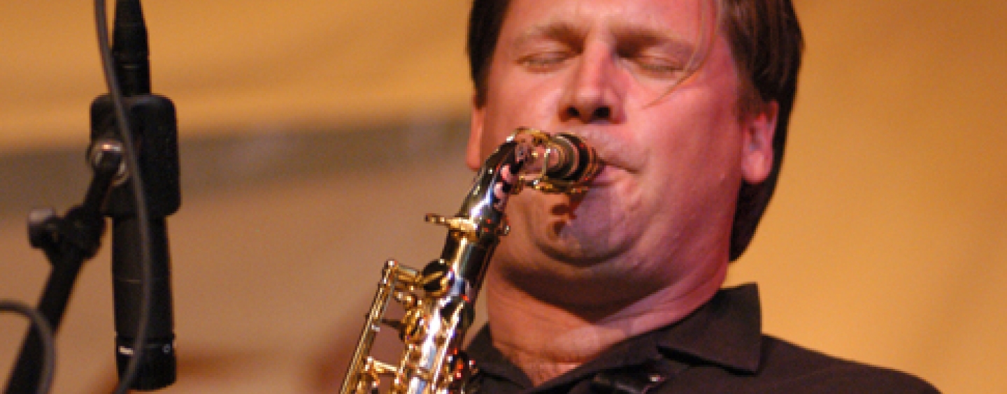 Andrew Speight, a white man in a black polo shirt, playing his saxophone. His eyes are closed and his hair pushed back from his forehead.