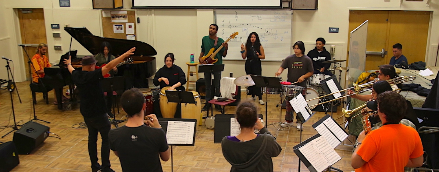 A group of students playing various wind, percussion, and stringed instruments rehearse in a circle in a classroom at SF State