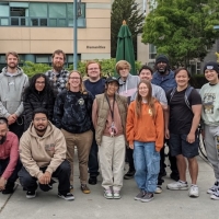 A group of students of diverse ethnicity and gender representation wearing casual clothing stand outside the Creative Arts building on SFSU campus. They are posed in rows and smiling at the camera.