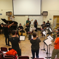 A group of students playing various wind, percussion, and stringed instruments rehearse in a circle in a classroom at SF State