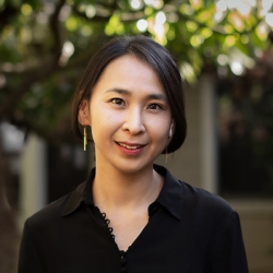 An Asian woman with chin-length dark hair wearing a black blouse with a v-neck stands in front of trees and buildings blurred in the background. 