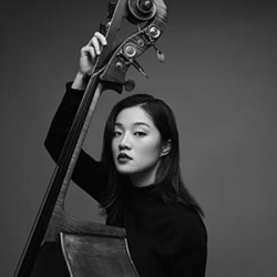 Yuchen Liu, a young Asian woman with shoulder-length dark hair and a black long-sleeved top looks at the camera and holds her Double-Bass against her body