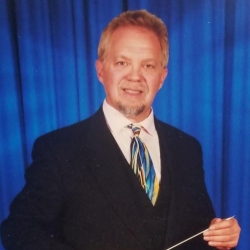 A white man wearing a dark suit and blue-striped tie looking at the camera and holding a conductor's baton in one hand