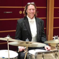 Allen Biggs, a white man with long blonde hair, seated behind a drum kit wearing a white tie tuxedo and smiling at the camera