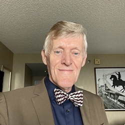 An older white man with short silver hair parted on the side wearing a drak shirt, brown jacket, and a bowtie
