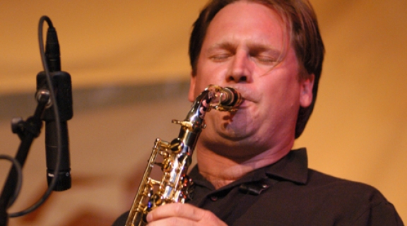 Andrew Speight, a white man in a black polo shirt, playing his saxophone. His eyes are closed and his hair pushed back from his forehead.