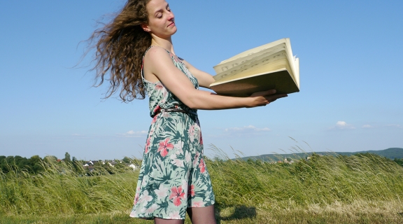 Johanna Tarscom, a young white woman in a floral sundress standing in a field holding open a large book of musicwearing